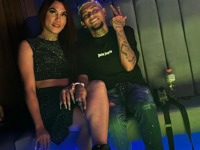 MegaSexyHot with dark hair in a sparkly black dress and MegaSexyHot with bleached hair, tattoos, and a black shirt making a peace sign, sitting together.