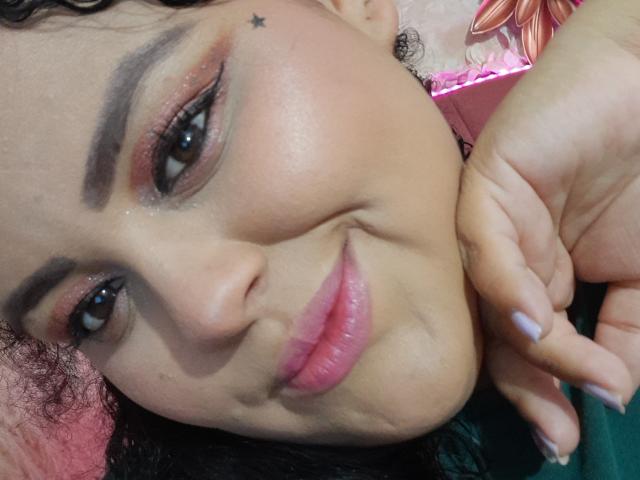 MelannieCarter's face, smiling with pink lips, brown eyes, and a small star tattoo above her right eyebrow. She has curly hair, dark eyebrows, and light pink nail polish.