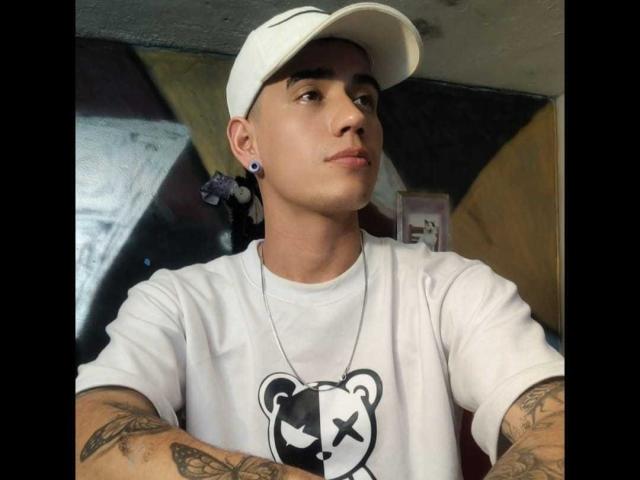 Jahnk is a young dude with a white cap, ear gauges, and tattoos, wearing a white shirt with a black bear graphic, looking serious with arms crossed.
