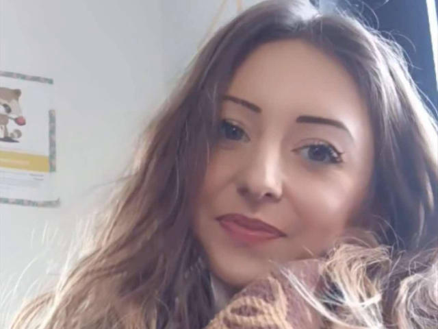 EmmaQueen69's face, with wavy brown hair, subtle makeup, and pink lipstick, smiling softly. Her tits are partially visible under a brown sweater. She has a cute, sexy look.