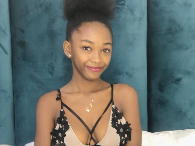 Jaurita with dark skin, small tits, in a lace bra, smiling, wearing a gold necklace, top bun hairstyle, blue velvet background, and a slight pink lip color.