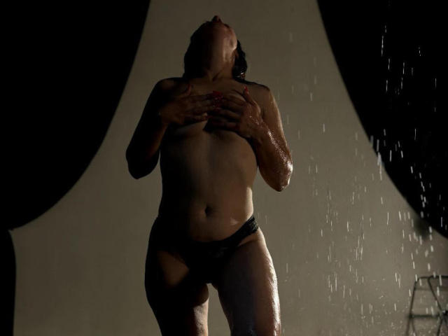 NikaAlvares stands under water, her wet skin glistening. She's topless, covering her tits with hands, wearing black panties. Water droplets fall around her, head tilted back.
