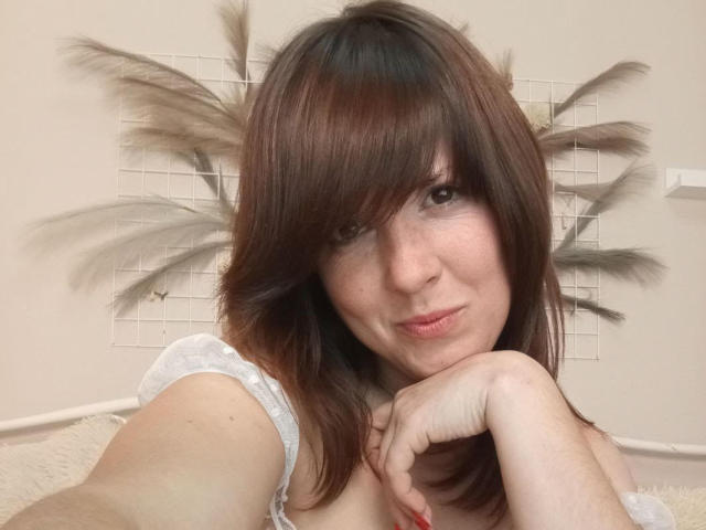 NikaAlvares with brown hair, pouty lips, and a cute face, resting her chin on her hand, showing her bare shoulders and white, puffy-sleeved top, and red nails.