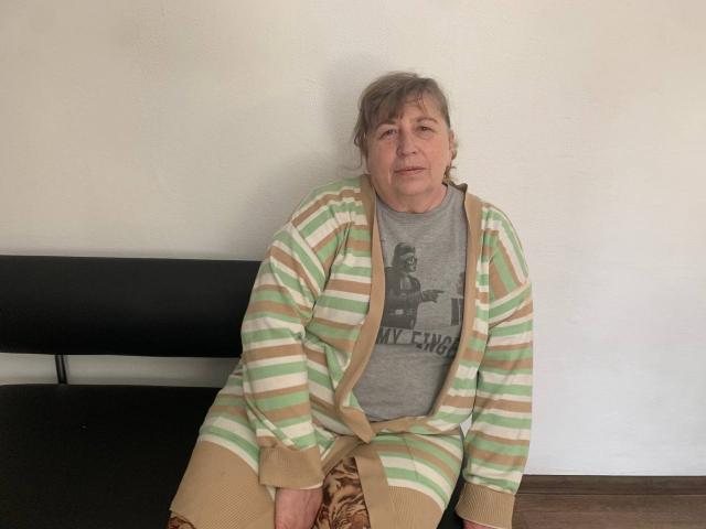 MagdalenaGlory with gray t-shirt, striped green and brown cardigan, sitting on black bench, gray hair, slight smile, wearing brown pants, hands on knee, casual, relaxed.
