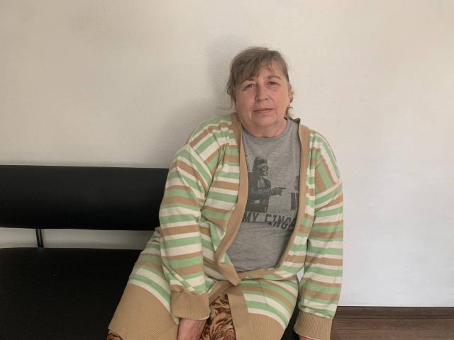 MagdalenaGlory, middle-aged woman with gray hair, wearing a green and beige striped cardigan over a gray shirt, sitting on a black bench, looking slightly tired.