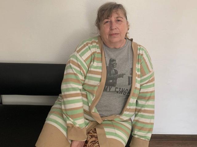 MagdalenaGlory is an old woman with gray hair, wearing a striped green, white, and beige cardigan over a gray shirt, sitting on a black bench.