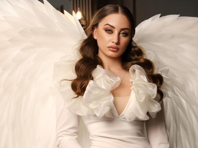 MaribelleVinnie with white angel wings, wearing a white dress with ruffled shoulders, has wavy brown hair, and a serious expression, showing her tits and a pretty face.