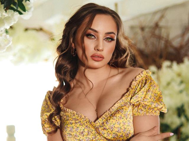 MaribelleVinnie with wavy brown hair, green eyes, and full lips, wearing a yellow floral dress, showing her tits, arms crossed, looking at the camera with a serious expression.