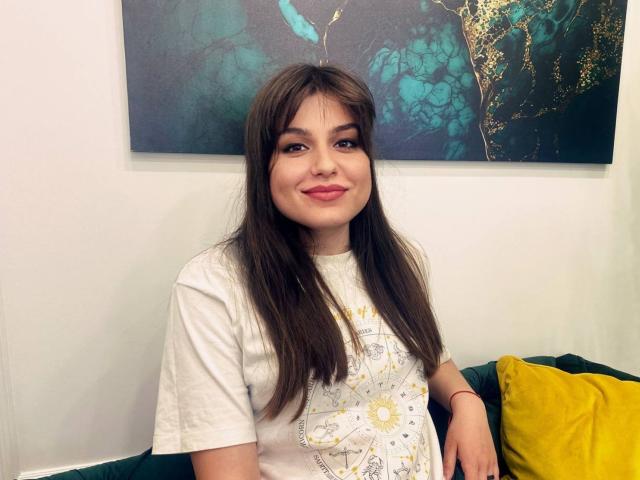 Amalia Potery, mit langem braunen Haar, einem weißen Hemd und pinkem Lippenstift, sitzt lächelnd auf einem grünen Sofa mit einem gelben Kissen, ihre Hand auf ihrem Oberschenkel..