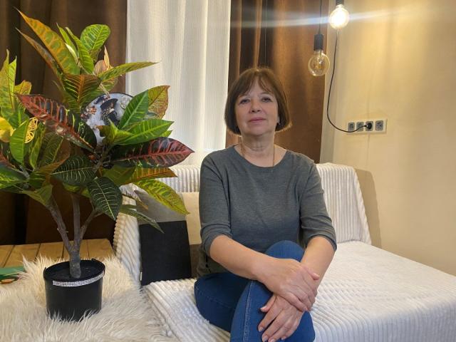 VanessaOrchid, une femme d'âge moyen avec des cheveux bruns courts, un t-shirt gris, un jean bleu, assise sur un canapé blanc, les mains sur les genoux, une petite plante en pot sur la gauche, une ampoule suspendue derrière..