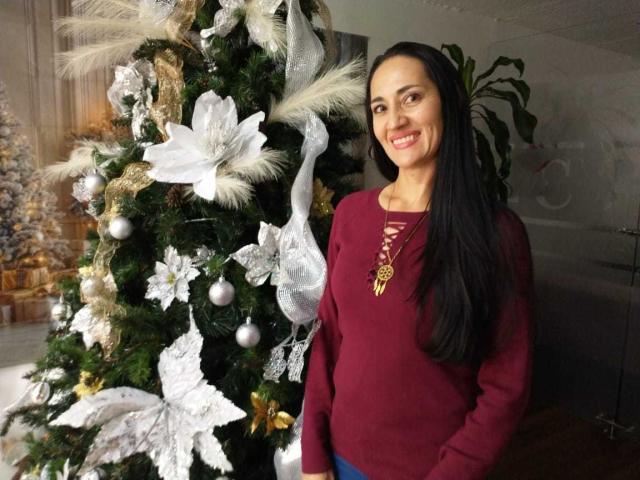 MiaMott with long black hair, smiling, wearing a red long-sleeve top, standing next to a decorated Christmas tree with white and silver ornaments. Her tits are small.