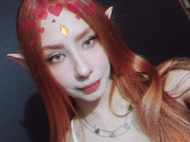 Spitfirex with red elf ears, green eyes, pale skin, and long red hair. Red heart stickers on forehead. Gold diamond ornament on forehead. Smiling. Necklace with small charms.