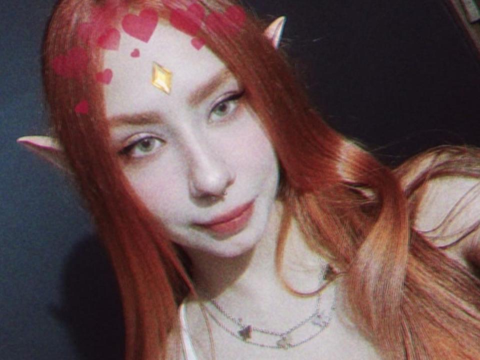 Spitfirex with red elf ears, green eyes, pale skin, and long red hair. Red heart stickers on forehead. Gold diamond ornament on forehead. Smiling. Necklace with small charms.