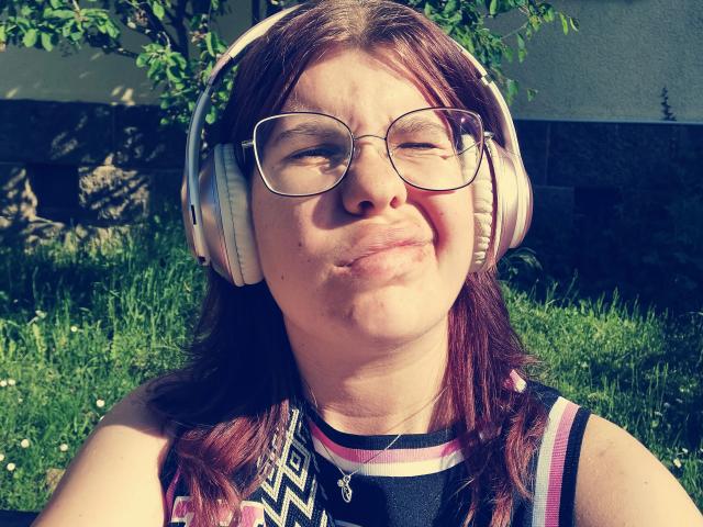 Tessa35, con cabello rojo, gafas y auriculares rosas, sonriendo con una nariz ligeramente inflamada, vestida con una camisa de color negro y rosa con estampado, sentada sobre hierba verde..