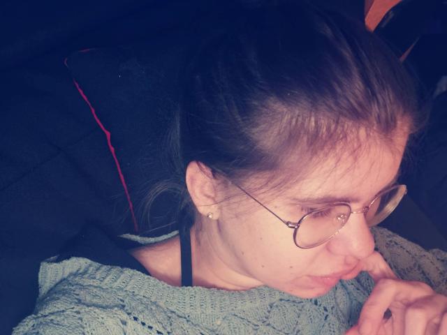 Tessa35: aspetto attraente, con occhiali, viso leggermente gonfio e capelli castani disordinati, indossa un maglione verde, leggermente piegata in avanti con un dito vicino alla bocca..