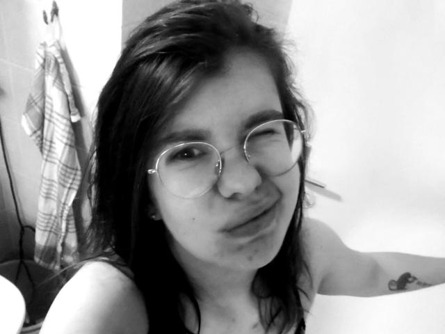 Selfie in bianco e nero di Tessa35-hot con capelli scuri ondulati, occhiali rotondi, un leggero sorriso, un naso piccolo e un tatuaggio visibile sul braccio destro, che indossa un top senza spalline..