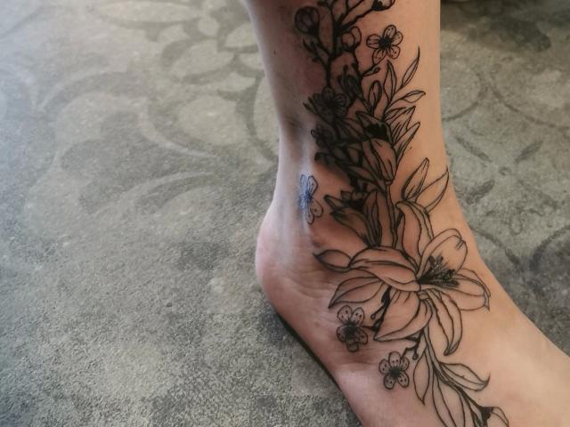 El tatuaje en el tobillo de Tessa35-hot presenta lirios, hojas y pequeñas flores dibujados con tinta negra detallada, que cubren la parte inferior de su pie y se extienden ligeramente por encima de su hueso del tobillo..