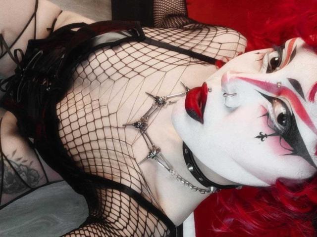 GothVioleth met witte schmink, rode lippenstift en rood haar, gekleed in een nethemd, zwart leer en een metalen ketting, waarbij haar borsten en tatoeages zichtbaar zijn, liggend..