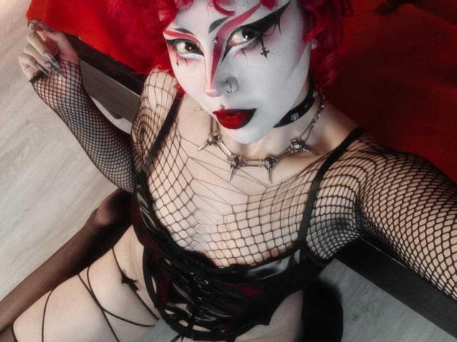 GothVioleth con cabello rizado rojo, maquillaje blanco en la cara, lápiz labial rojo, top de malla negra, corpiño negro, collar de cadena, sentada en el suelo, mostrando sus pechos y tirantes negros..