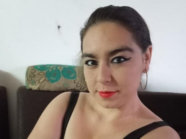 A TaylorNova tem cabelo escuro, delineador preto, batom vermelho, brincos de argola e um piercing no nariz. Ela está vestindo uma blusa preta com alças, que mostra seus seios. Ela está olhando para a câmera..