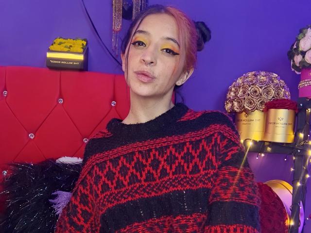 VanelopeSmith with red and black patterned sweater, yellow eye makeup, pink hair in bun, pouty lips, sitting on red tufted bed, surrounded by decorative items, purple wall.