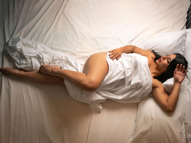 AliceFoster69, incinta e con un sedere prosperoso, giace nuda sotto un lenzuolo bianco, coprendo i seni e i genitali, con la mano destra sul ventre e la mano sinistra sopra la testa..