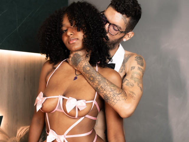 Jenifer et Benn, avec leurs cheveux noirs bouclés, portent de la lingerie rose à bretelles avec des nœuds qui couvrent leurs seins. Jenifer se tient devant Benn, qui a des tatouages, porte des lunettes, un nœud papillon noir et une chemise noire..