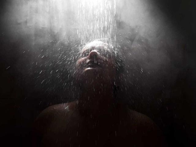 Le visage de PratanoHot est illuminé par l'eau qui tombe, la tête penchée en arrière, l'eau ruisselant, les yeux fermés, la bouche légèrement ouverte, arrière-plan sombre et brumeux, torse nu, gouttes d'eau visibles..