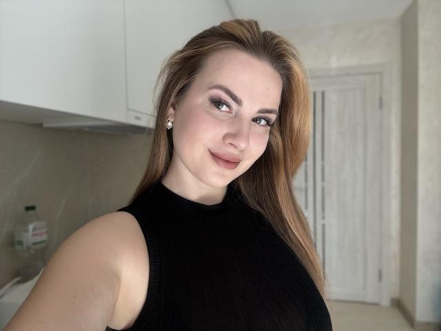 MissTemptationI with light brown hair, wearing a black sleeveless top, smiling at the camera, showing her boobs, in a kitchen with white cabinets and a door in the background.