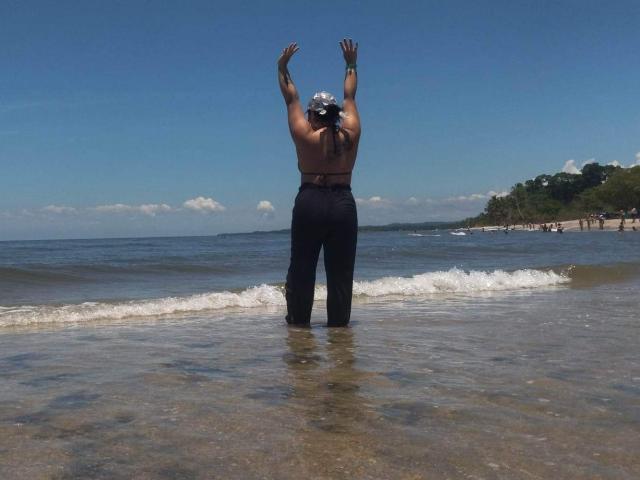 ValentinaSilva stands in shallow water, arms raised, shirtless with black pants, tattoos on arms, cap on head, facing ocean, clear blue sky, small waves, green hill in background.