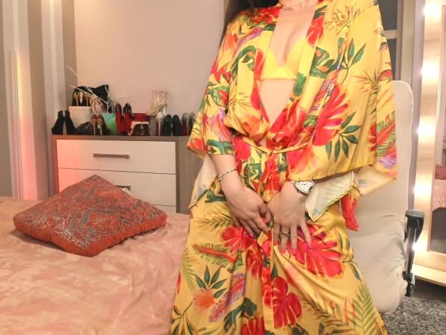 LinaLuchia wearing a yellow floral robe with red flowers, exposing her yellow bra-covered tits, hands on her robe, white chair, pink bed with orange pillow, and dresser in background.