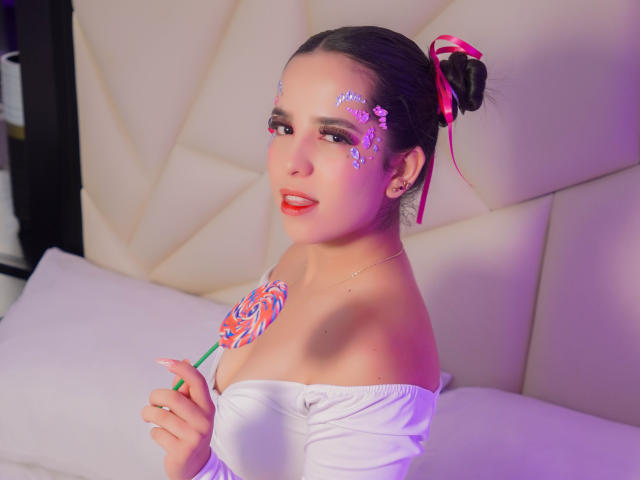 AlanaReyes with purple face glitter, holding a pink and blue swirl lollipop, wearing an off-shoulder white top, pink hair ribbon, and earrings, smiling with red lipstick.