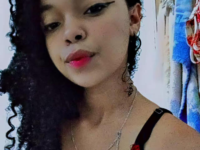 LeilaSweety with curly black hair, pink lipstick, and black strap dress, showing a cute, confident face, wearing a thin necklace, with a subtle smile and slight nose ring.