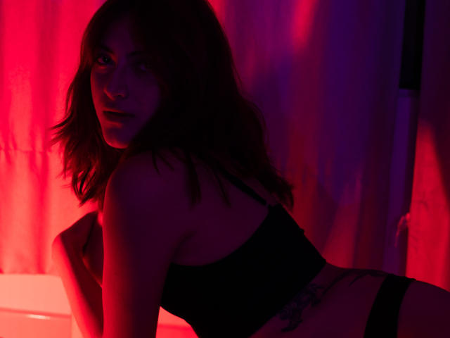 SpencerLe with dark hair, wearing a black bra and black panties, leaning forward in red and purple lighting, showing a tattoo on her side, looking at the camera.