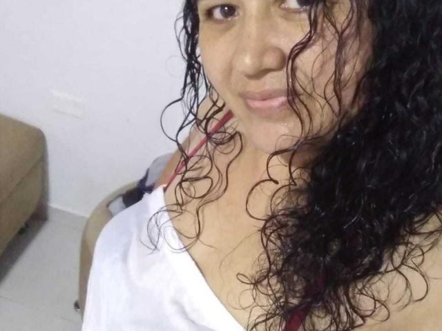 Kleimar has curly black hair, brown eyes, and a small smile. She's wearing a white shirt, exposing her shoulder and a red strap. Boobs are partially visible.