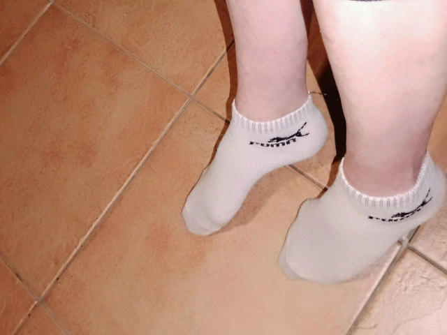 Madamecatsyve-hot's white socks, slightly dirty at the bottom, are on her pale legs standing on orange-tiled floor. Her toes are visible, and the socks have a small black logo.