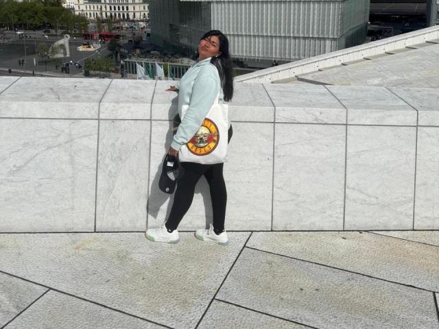 IvySoul with black hair, light blue hoodie, black leggings, white sneakers, holding bag with yellow cartoon, leaning against white marble wall, smiling, standing on gray textured pavement.