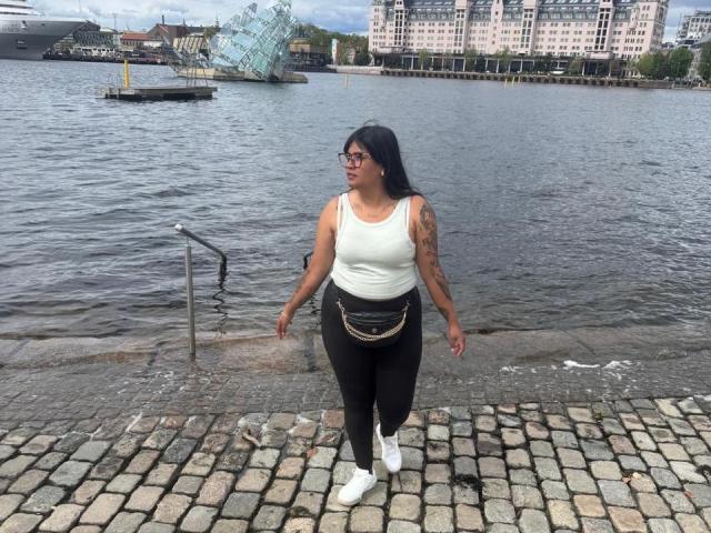 IvySoul has dark hair, glasses, white tank top, black pants, white sneakers, and a rope belt. She's walking on cobblestone by a waterway with a glass sculpture.