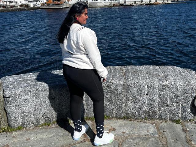 IvySoul tiene unas curvas pronunciadas, lleva leggings negros, zapatillas blancas y una sudadera con capucha blanca, y está de pie junto a una pared de piedra con agua azul en el fondo..