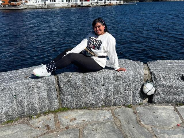 IvySoul is sitting on a stone wall, wearing black pants, white sneakers, and a white hoodie, with a black and white patterned belt, smiling at the camera.