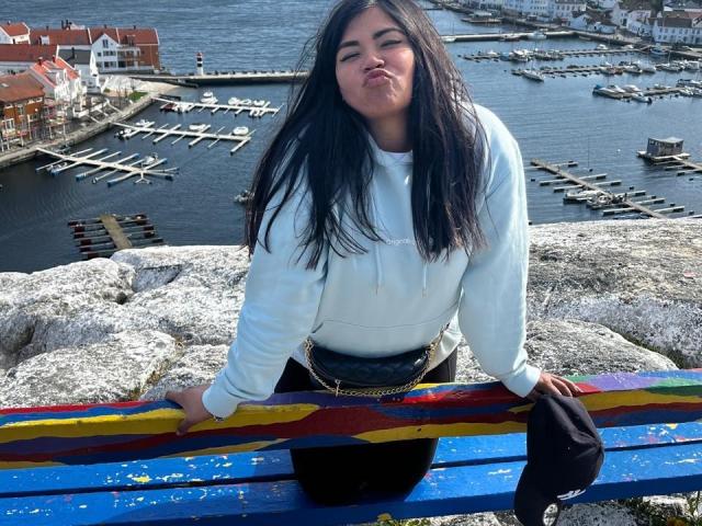 IvySoul with long black hair, white hoodie, black purse, and black pants, pouting at a colorful painted bench, holding a black cap, standing on rocky terrain.