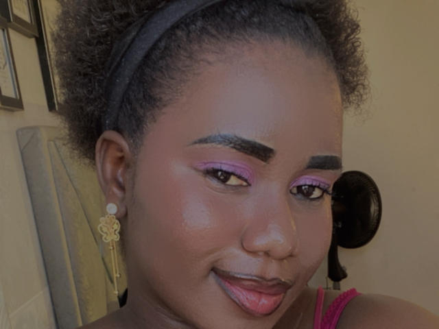 Dilay has dark skin, curly black hair, purple eyeshadow, thick eyebrows, gold earrings, and is sticking her tongue out. Her pink lipstick is slightly visible.
