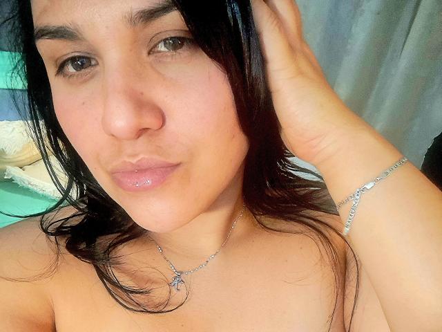 VivianaThompson has shiny pink lips, dark brown hair, and a silver necklace with a circular pendant. She's touching her hair with a silver bracelet on her wrist.