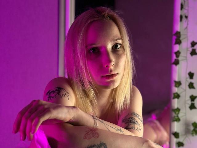 EllaXWilson, seminua com cabelo loiro, luz neon rosa, tatuagens nos braços e no peito, braços cruzados, olhando para a câmera, luz rosa no rosto, lábios ligeiramente entreabertos, expressão relaxada..