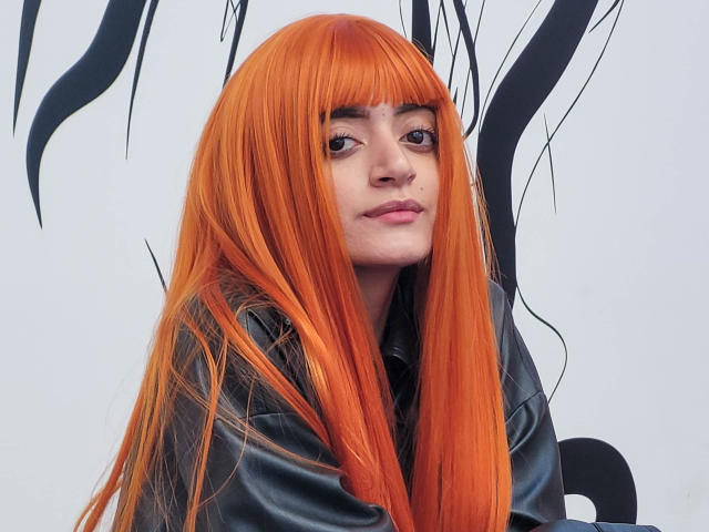 Amelie Sato mit langen, leuchtend orangefarbenen Haaren, die eine schwarze Lederjacke trägt, blickt leicht zur Seite mit einem neutralen Ausdruck, hat helle Haut und dezentes Make-up..
