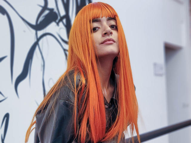 Amelie Sato mit leuchtend orangefarbenem, glattem, schulterlangem Haar, trägt eine schwarze Lederjacke, blickt leicht lächelnd nach oben und ihre Hände ruhen auf einem Geländer..