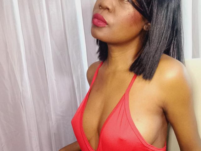 Karhinna with medium brown skin wearing a red bikini top, showing her small tits and shiny skin. She has straight black hair, red lipstick, and a nose ring.