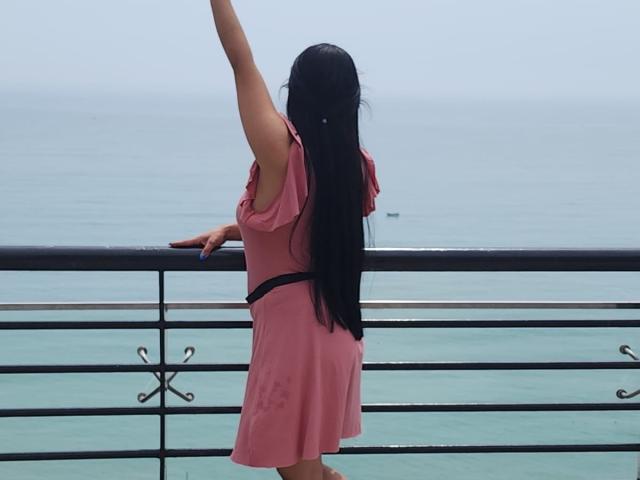 HeraDiosa with long black hair, pink dress, black belt, raising arm, standing on balcony, facing ocean, hand on railing, small boat in distance, calm blue water.