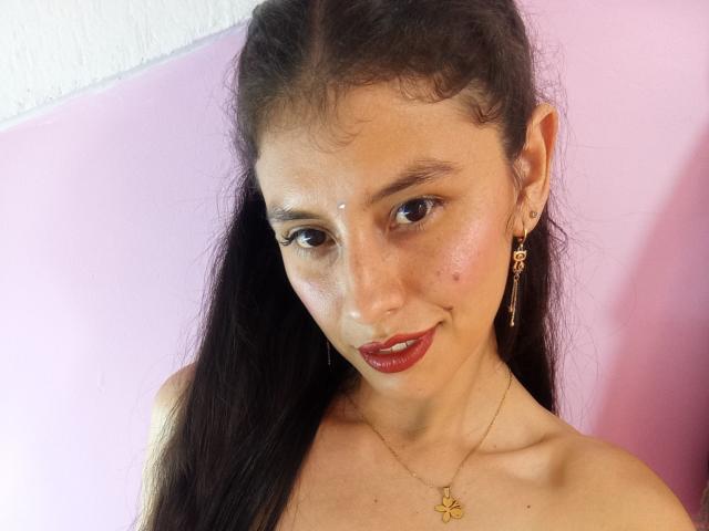 MadissonFontaine has long black hair, brown eyes, red lipstick, a gold necklace with a butterfly, and earrings. Her face is slightly freckled, with a small white dot on her forehead.