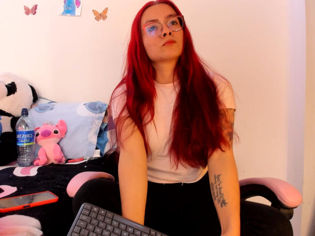 EimmyHoskX with long red hair, glasses, white t-shirt, black pants, tattoo on right arm, sitting on pink and black chair, water bottle, pink plush toy, keyboard in front.